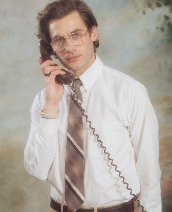 A man wearing glasses, a white shirt, and a striped tie is holding a corded telephone to his ear, standing in front of a mottled backdrop.