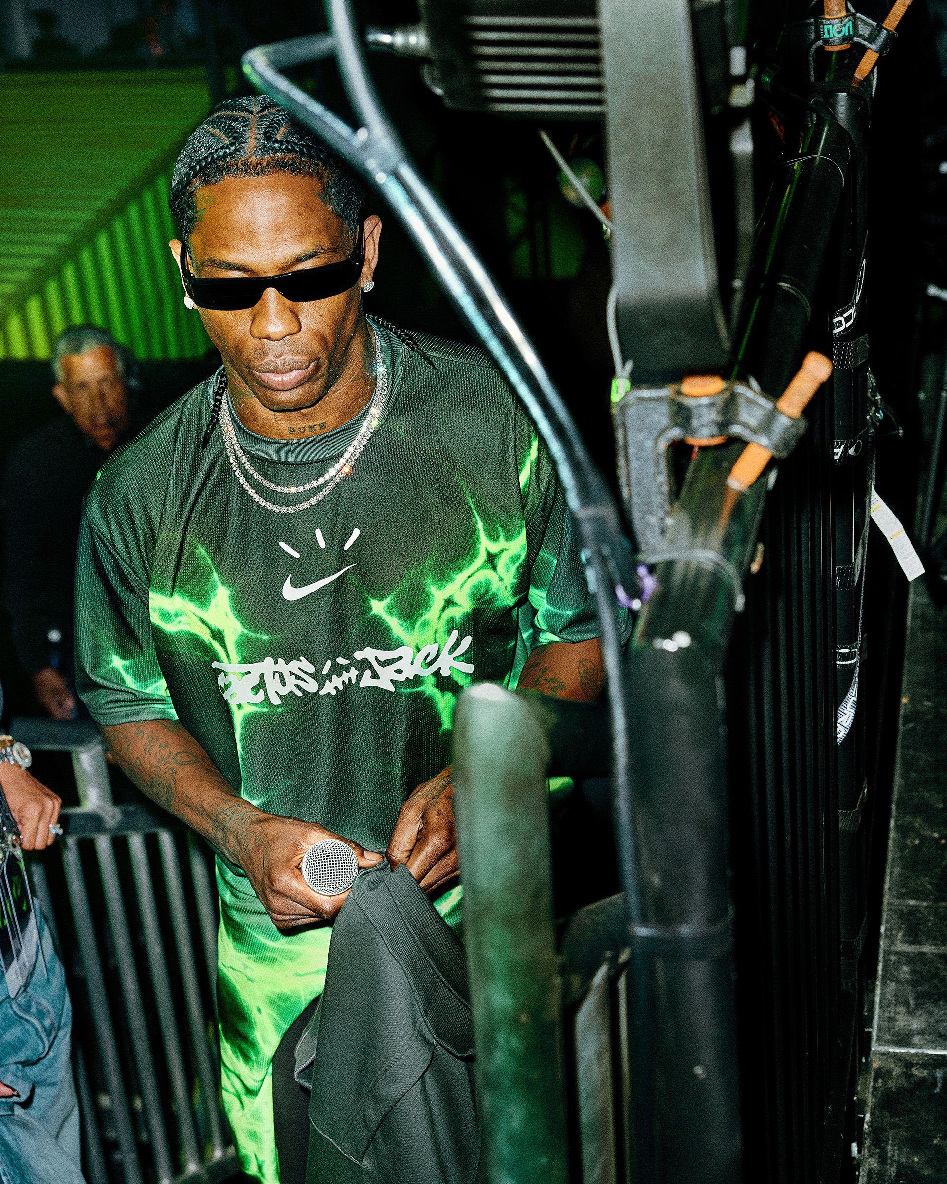 A man wearing sunglasses and a black and green shirt holds a microphone and a jacket while standing next to stage equipment.