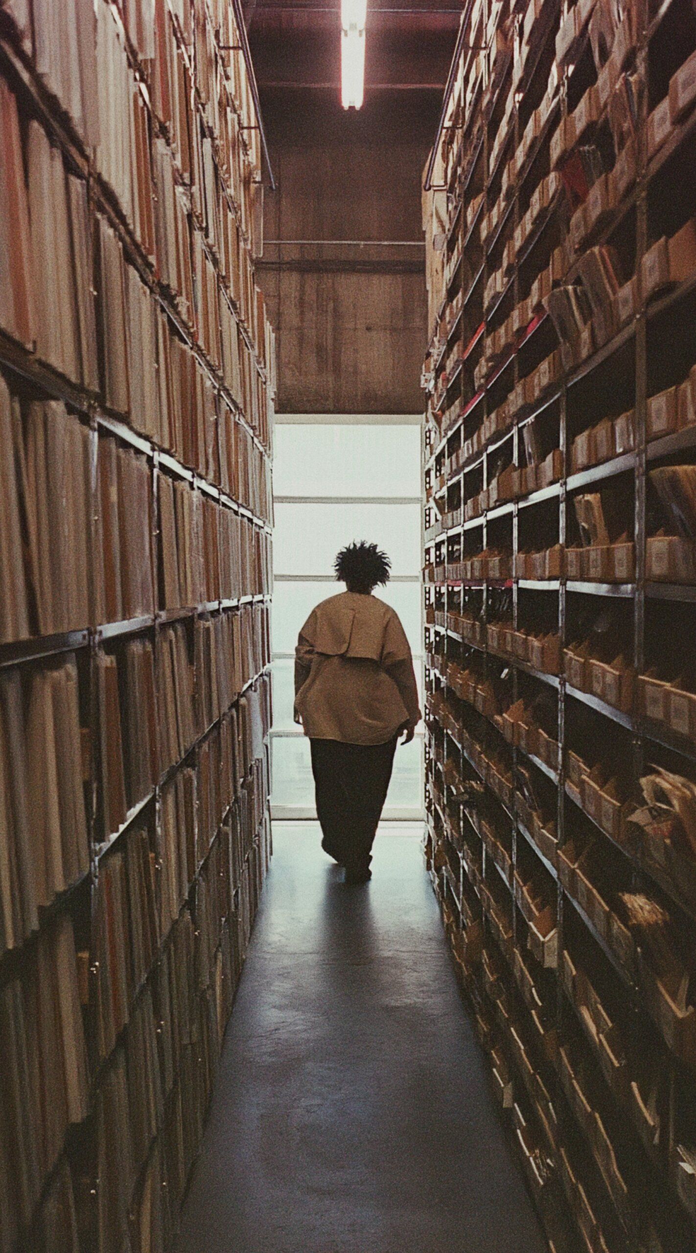 A person walks down a narrow aisle between tall shelves filled with archived files and documents in a dimly lit storage area.