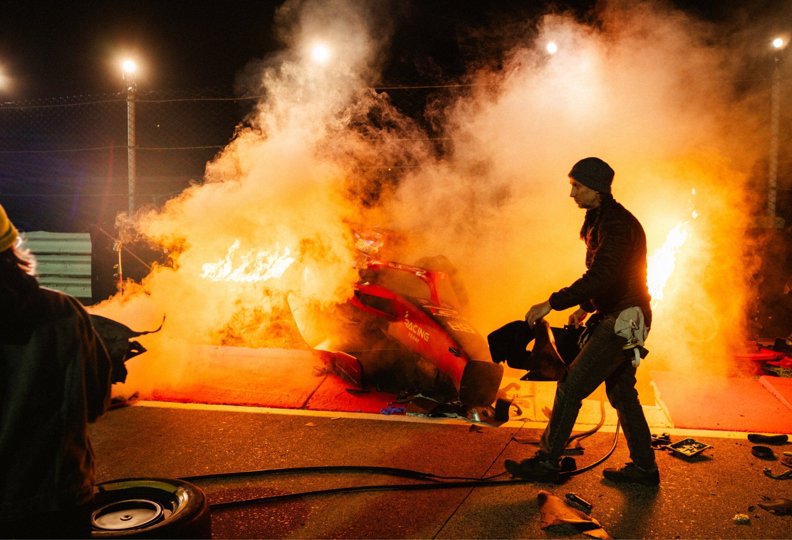 Person walking near a burning vehicle at night, with flames and smoke rising. Several lights illuminate the scene, and debris is scattered on the ground.