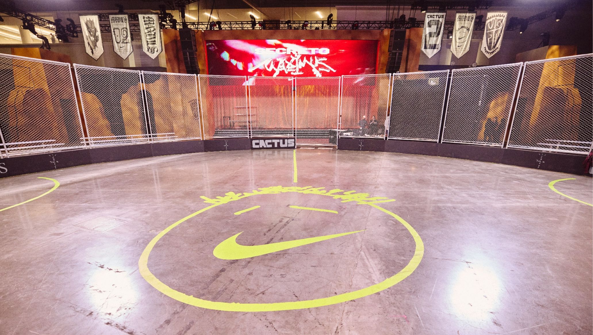 Indoor sports arena with fenced perimeter, a large neon yellow smiley face on the floor, banners hanging above, and a large screen at the back displaying the word "CACTUS.