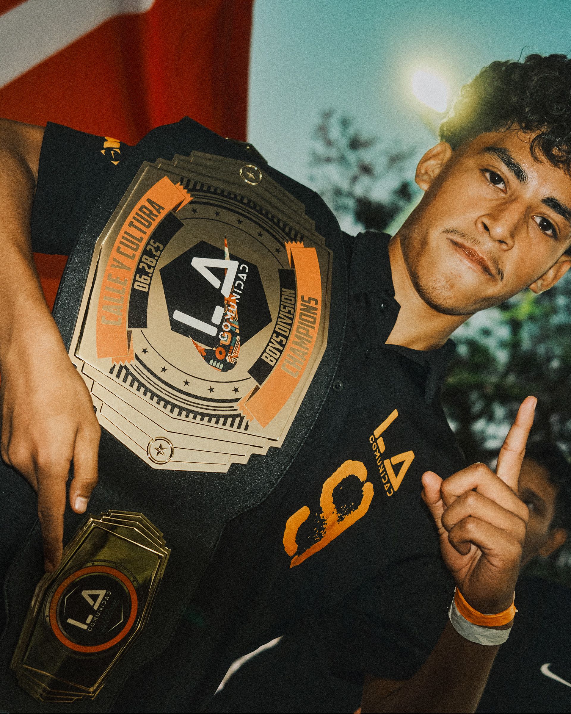 A young man holds a large championship belt over his shoulder and raises one finger, standing outdoors with a flag in the background.