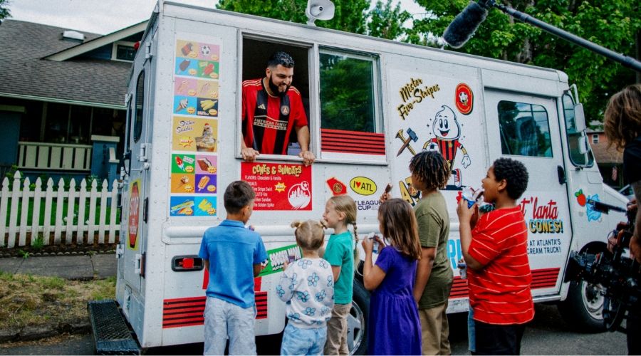 A man in a sports jersey serves snacks from a truck labeled "Mike's Hot Dogs" to a group of children who are standing in line outside. There is a person using a boom mic and another person taking a photo.