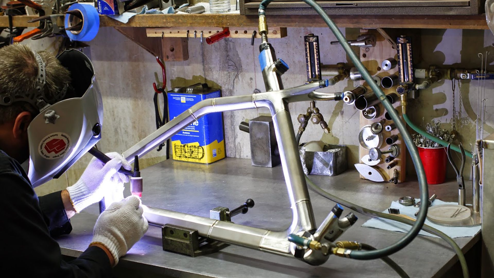 A person welding a metal bicycle frame in a workshop, surrounded by tools and equipment.