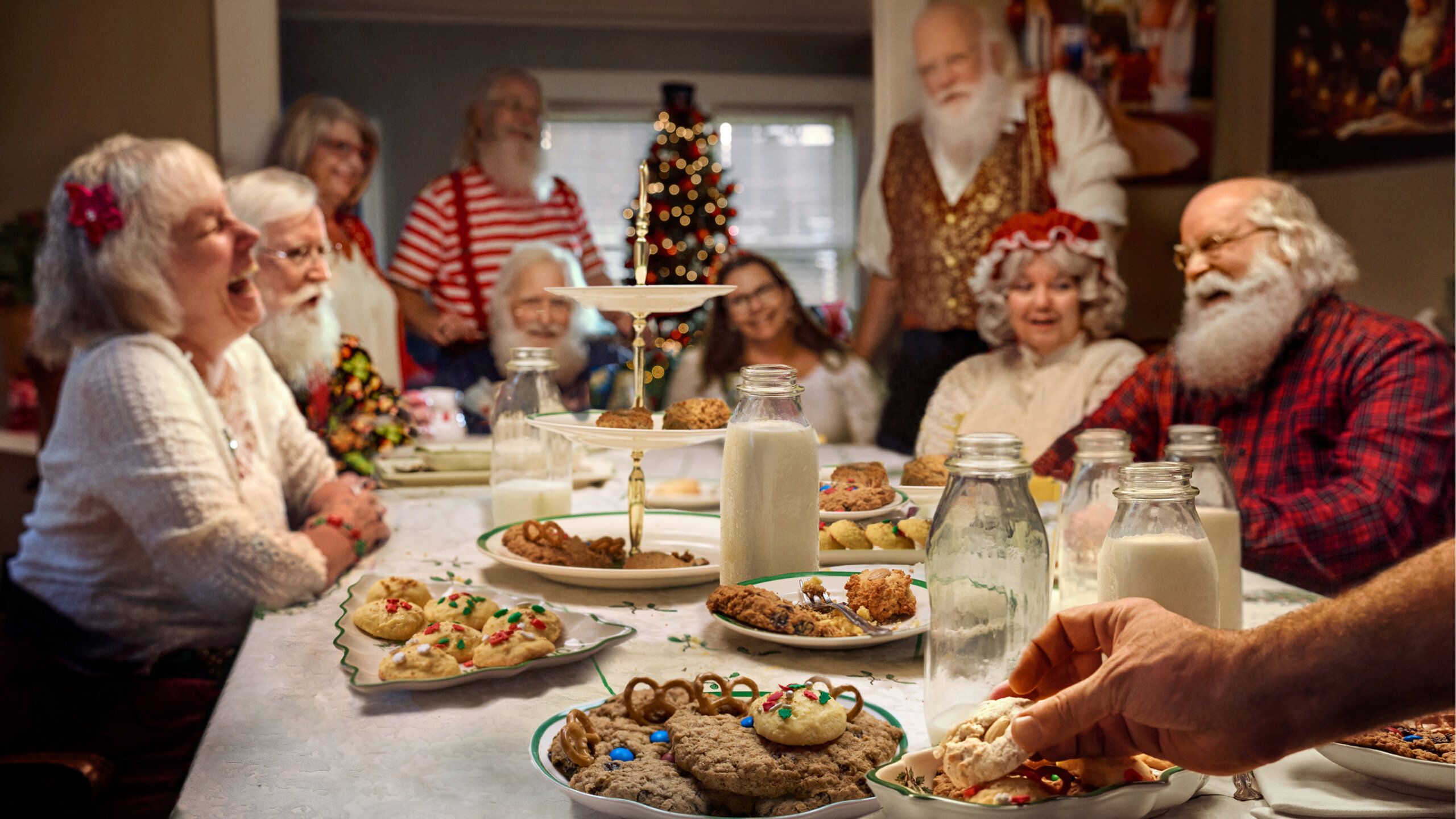 A group of elderly people, some dressed as Santa, sit around a table with cookies and milk, with a decorated Christmas tree in the background.