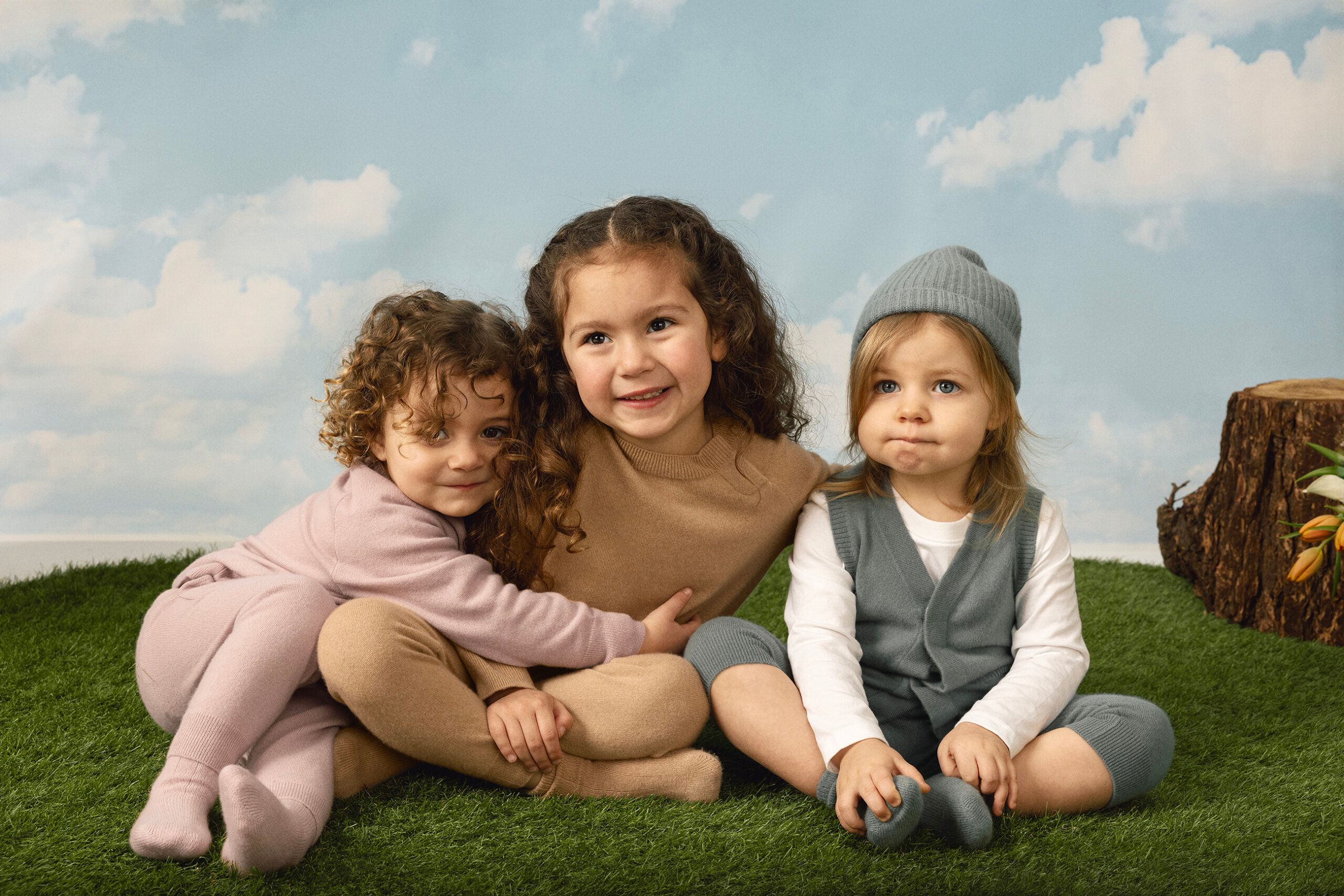 Three young children sit on fake grass in front of a sky backdrop. One child hugs another, while the third sits nearby wearing a blue hat. All are wearing soft, neutral-toned outfits.