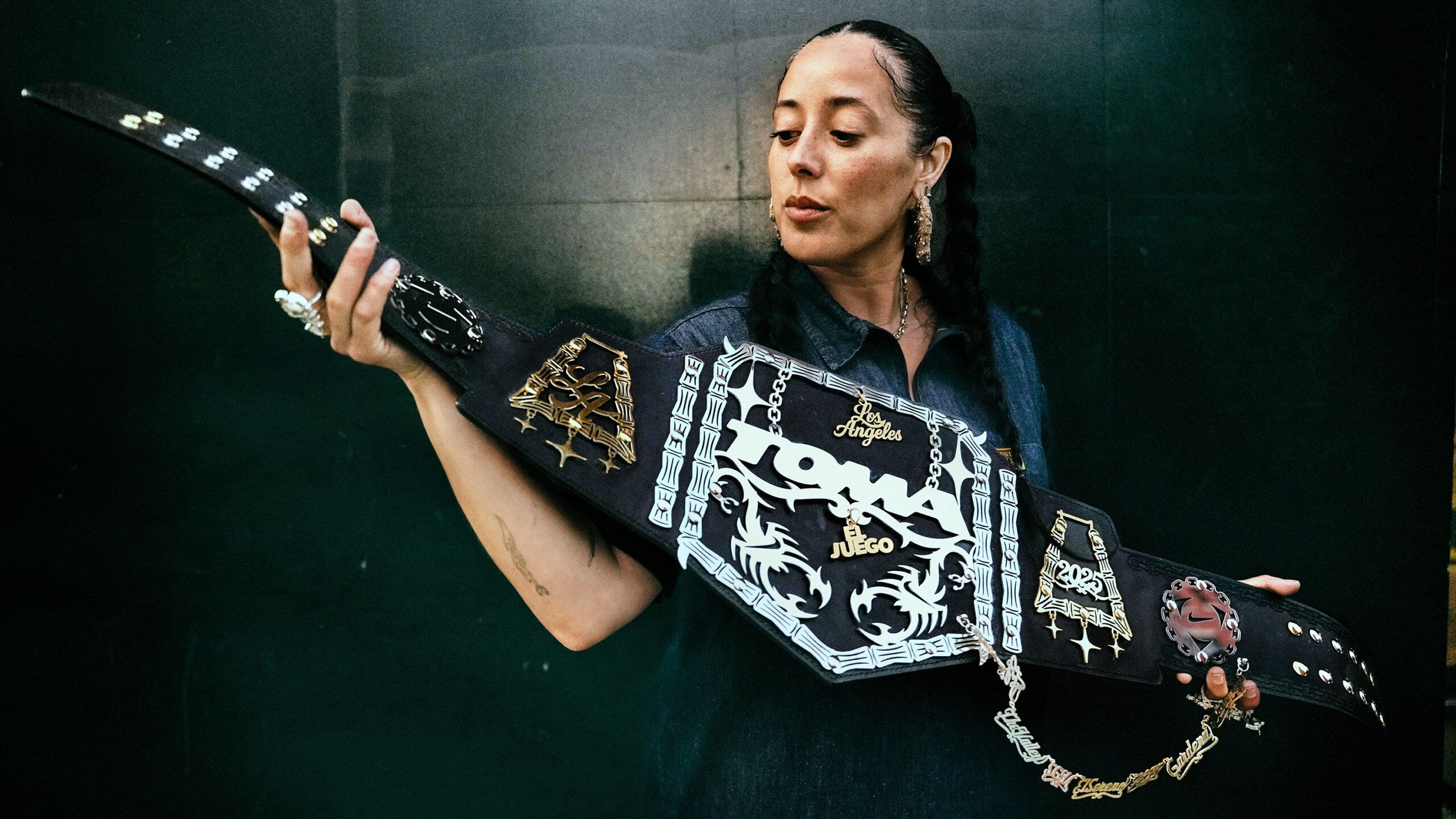 A woman in a dark shirt holds and examines a large, decorative championship belt against a dark green background.