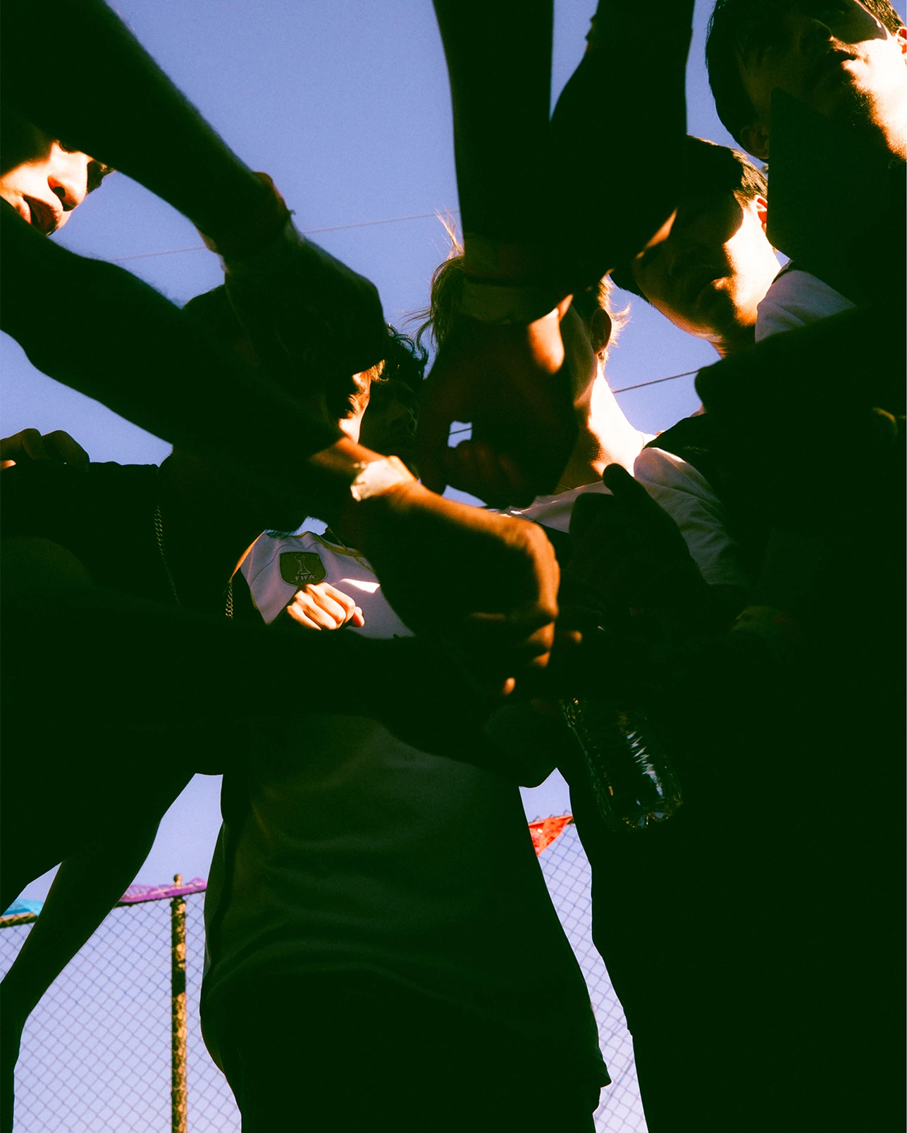 A group of people stand in a circle with their hands joined in the center, viewed from below against a clear sky.