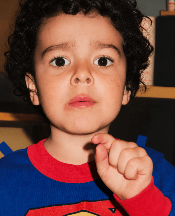 A young child with curly hair wearing a red and blue Superman shirt looks directly at the camera, holding their hand up near their face.