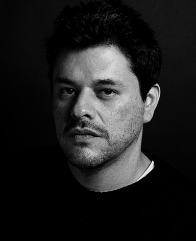 A man with short dark hair and facial stubble looks at the camera with a neutral expression against a dark background.