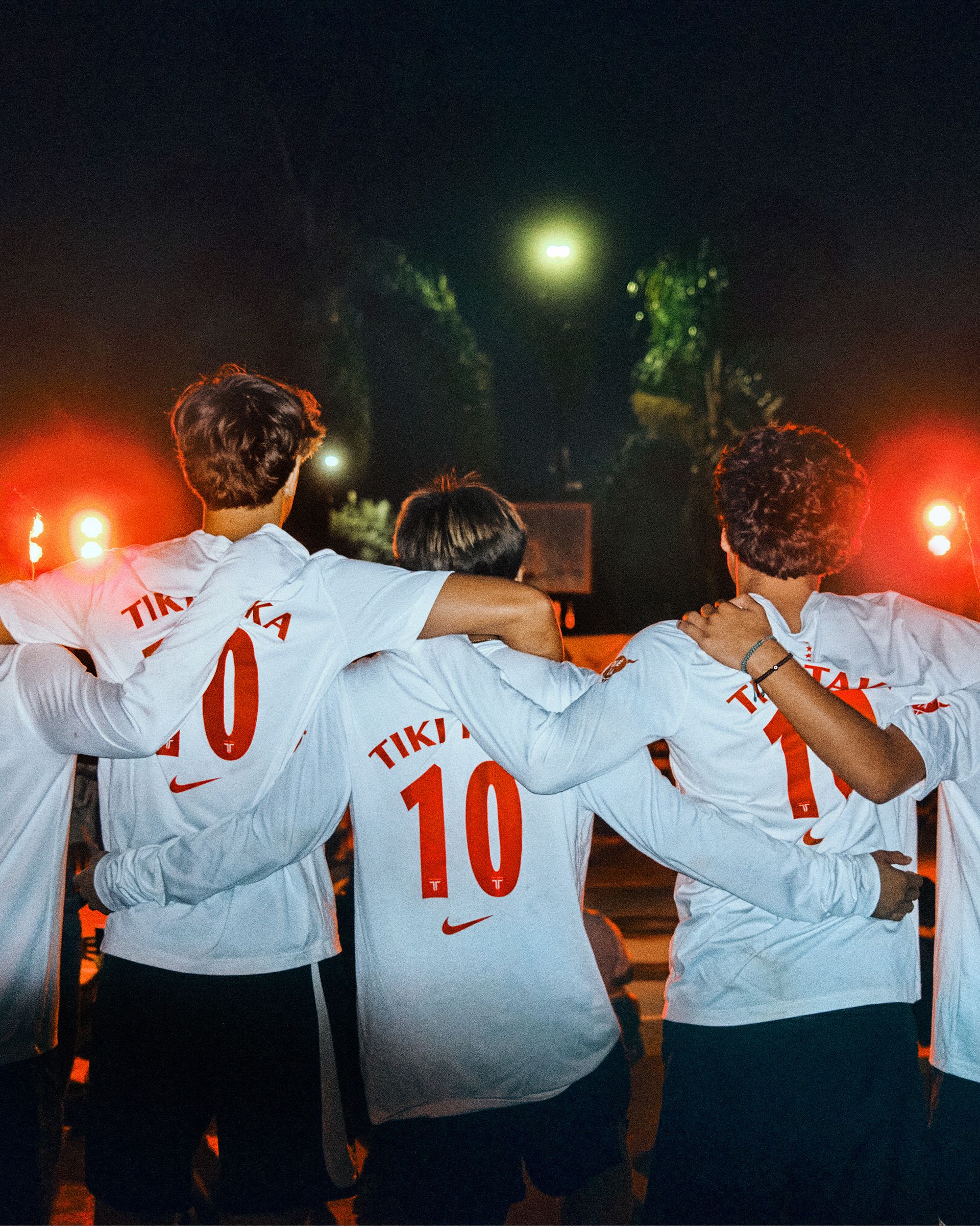 Four people wearing white soccer jerseys with the numbers 10 and 11 visible stand with arms around each other at night, facing away from the camera under bright lights.