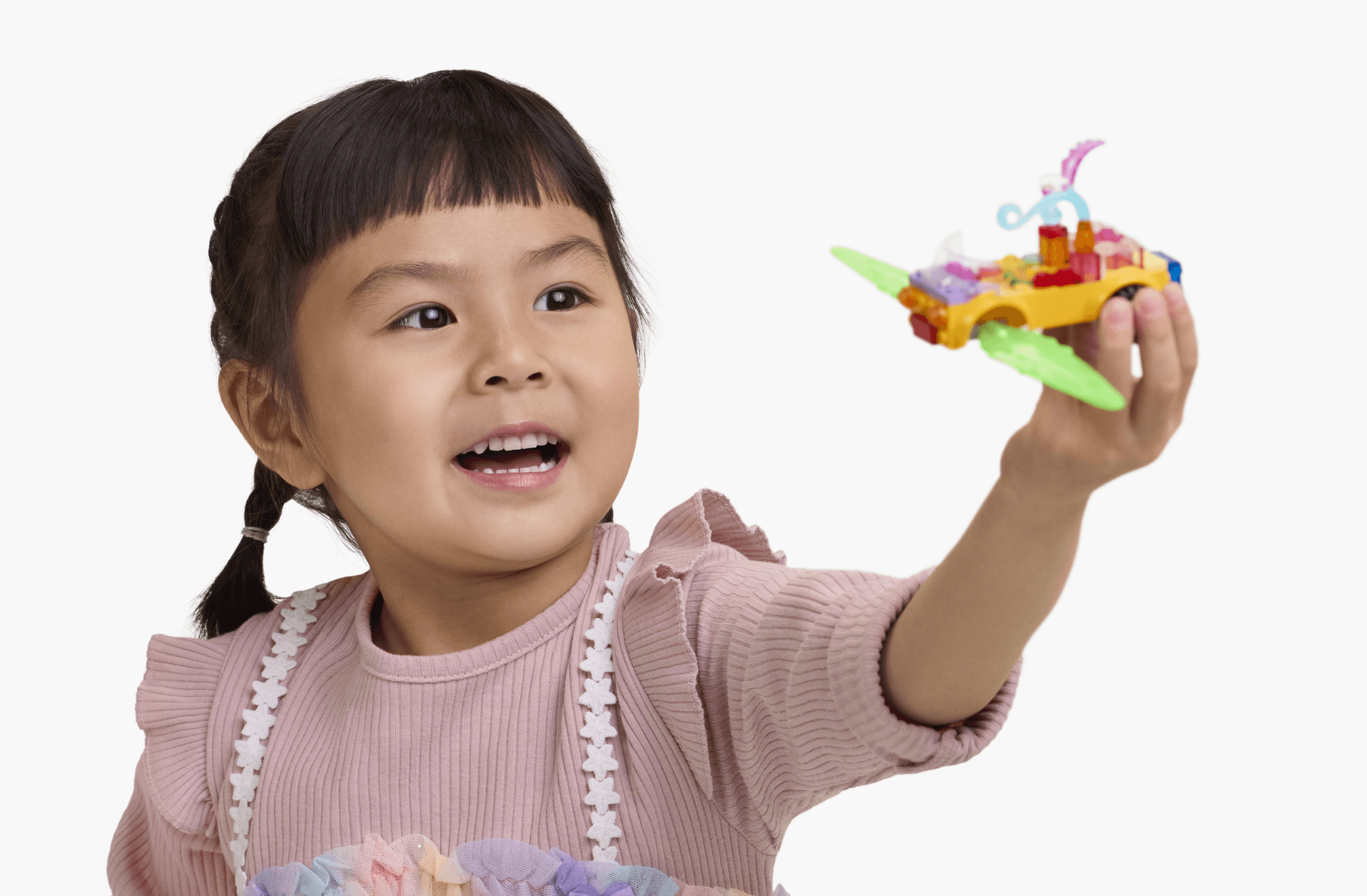 A young girl holds up a colorful toy plane, smiling and looking slightly to the side. She wears a pink shirt with frilled sleeves and has her hair in two braids.