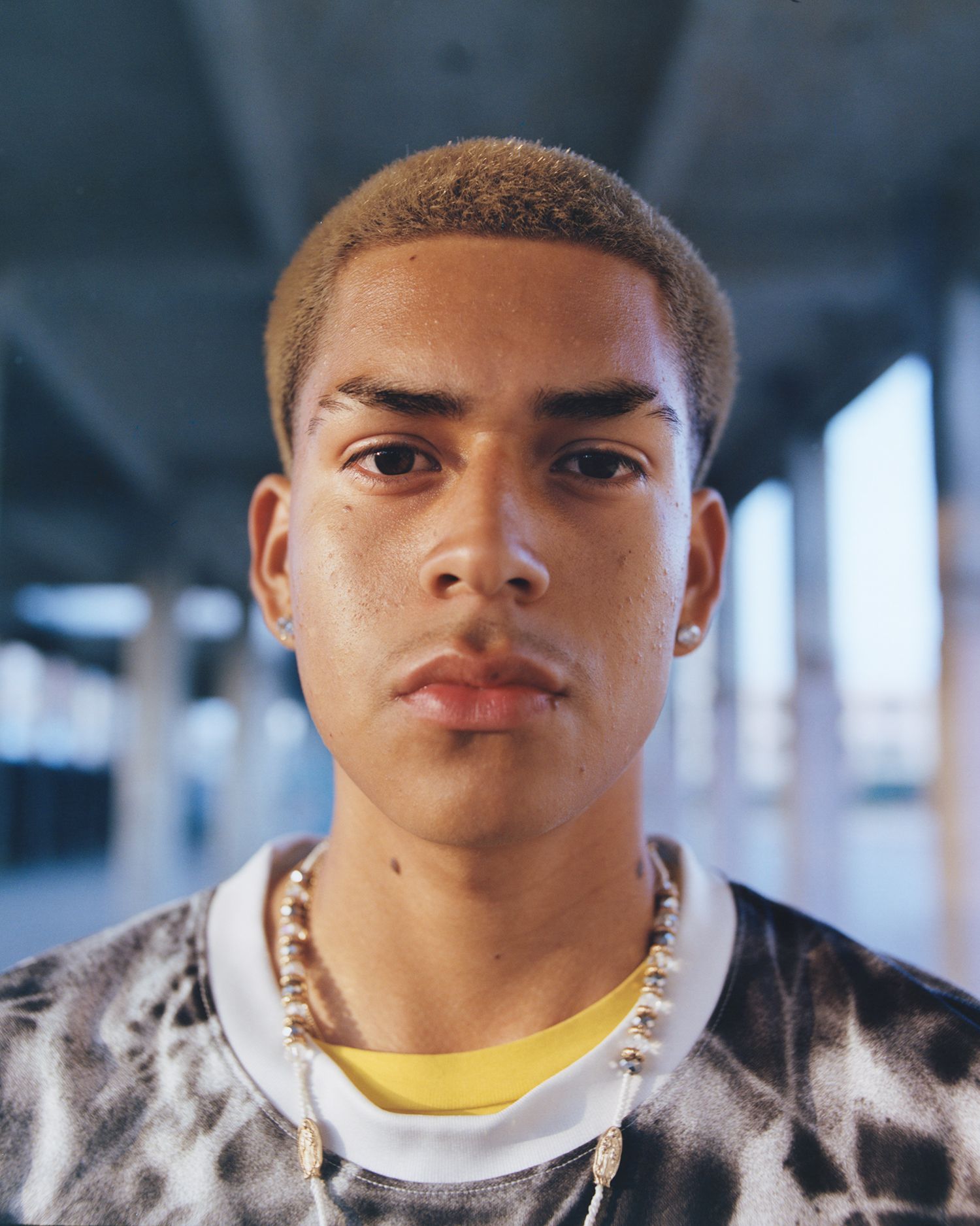 A young person with short, light brown hair, wearing earrings, a beaded necklace, and a tie-dye shirt, stands in an outdoor urban setting with concrete columns.