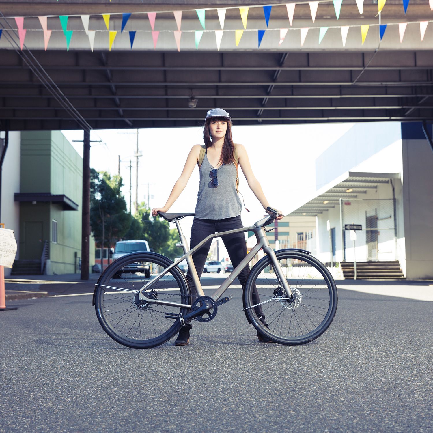 A young woman stands with her 3D printed bicycle under an overpass, wearing a helmet and casual clothing, with a street and small buildings in the background.