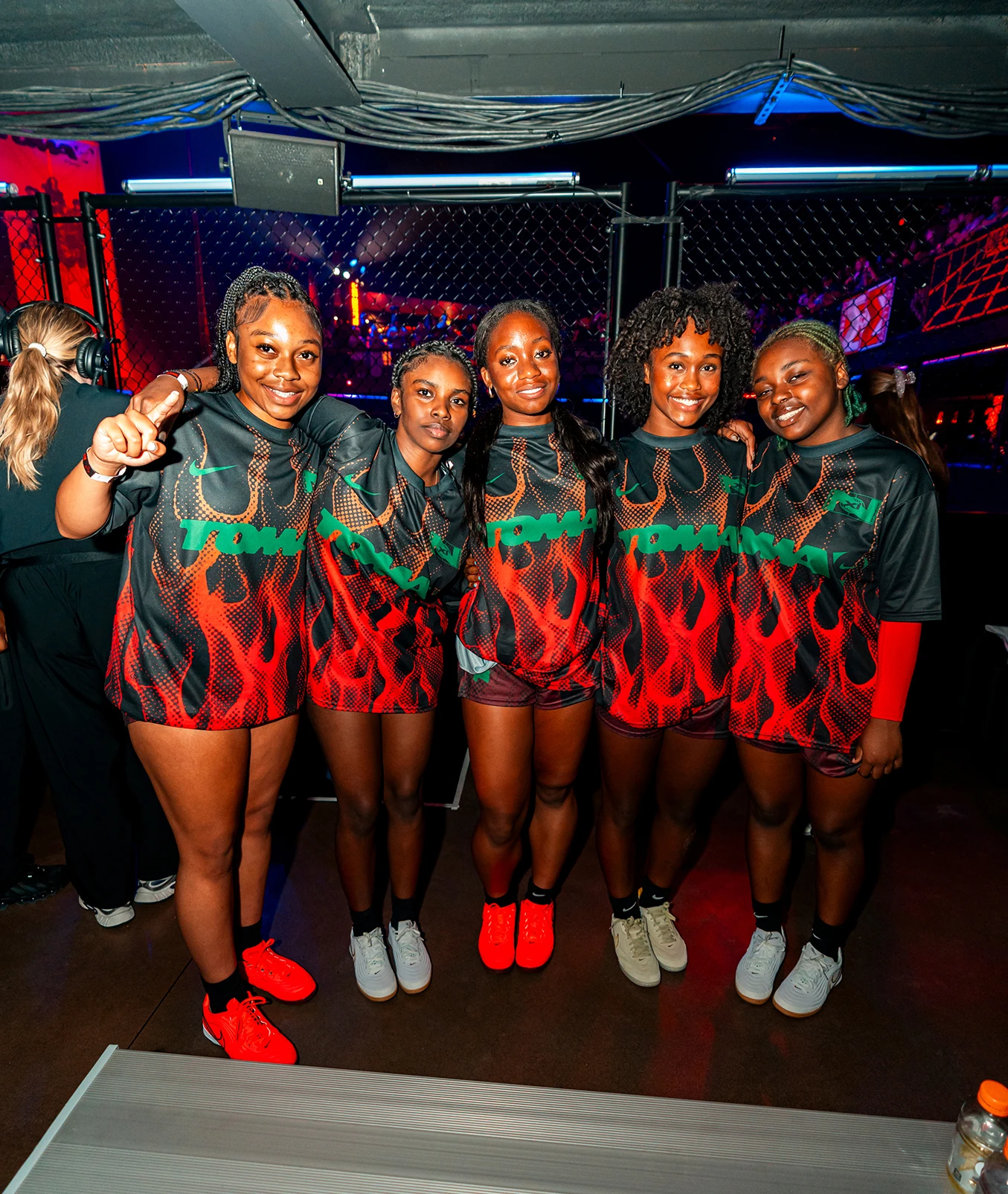 Five women in matching red and black sports uniforms stand side by side, smiling at the camera inside an indoor venue with dim lighting and a chain-link fence in the background.