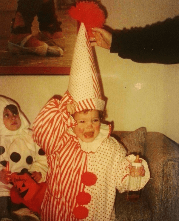 A young child in a red and white clown costume cries while holding a bottle. Another child in a polka dot outfit sits nearby. An adult adjusts the clown hat.