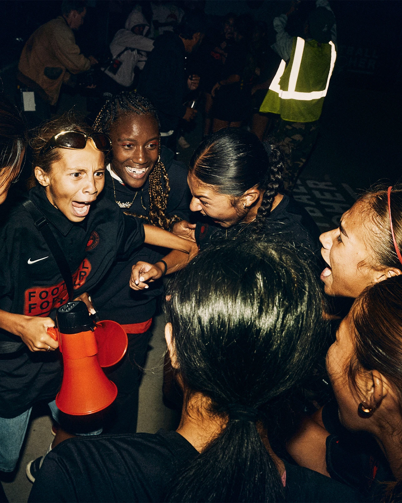 A group of women stand close together, some smiling and laughing, one holding a red megaphone. They appear to be celebrating at an event, with another person in a reflective vest in the background.
