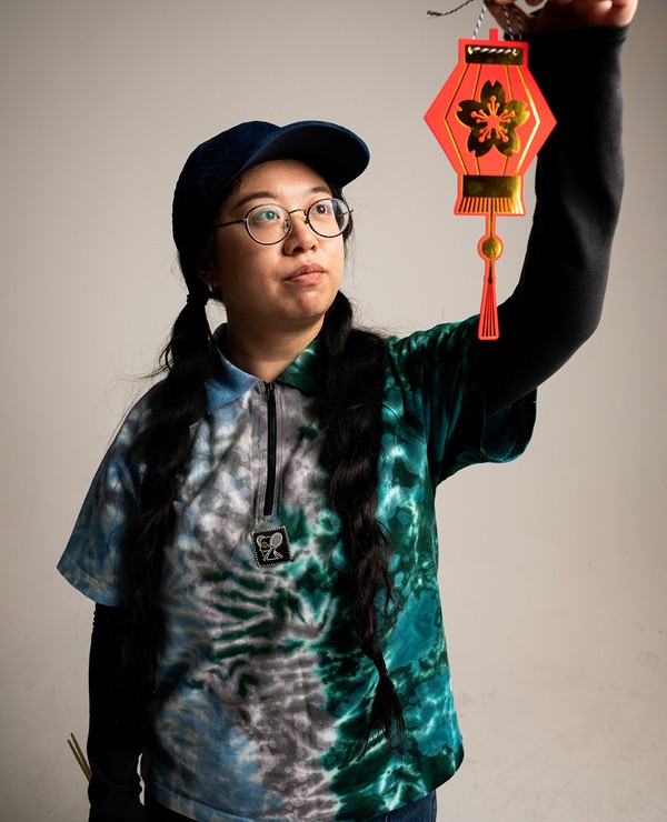 Person wearing glasses and a cap holds up a red and gold decorative lantern, looking at it closely. They have long braided hair and are dressed in a tie-dye shirt.