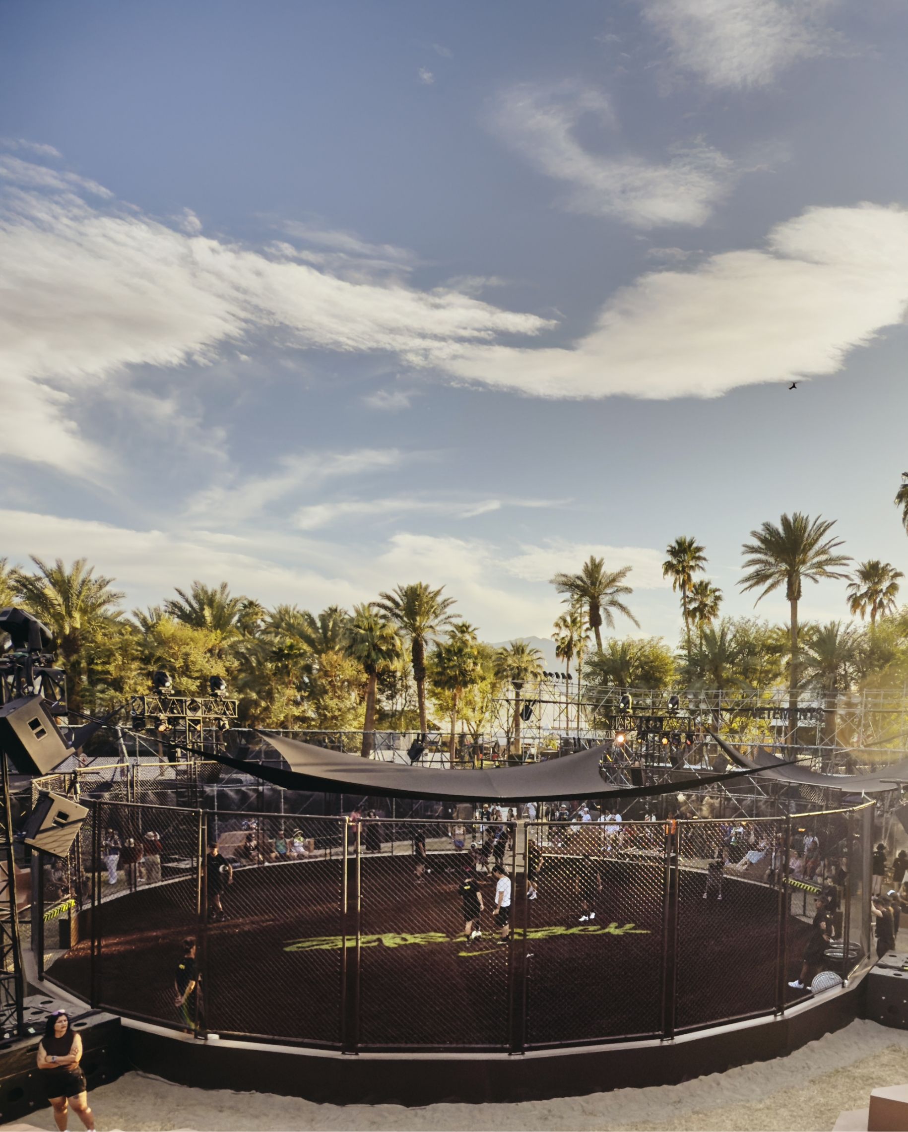 An outdoor sports court with a black mesh fence is surrounded by palm trees and stage lights under a partly cloudy sky.