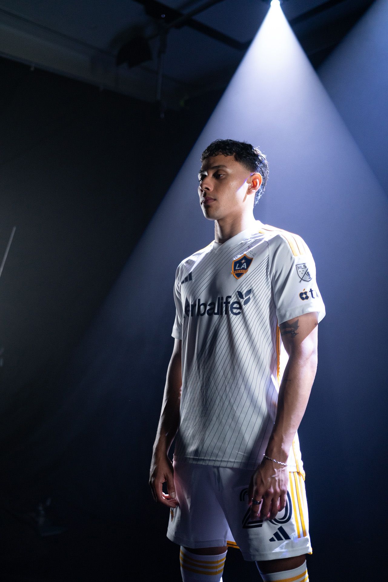 A soccer player in an LA Galaxy uniform stands under a spotlight against a dark background.