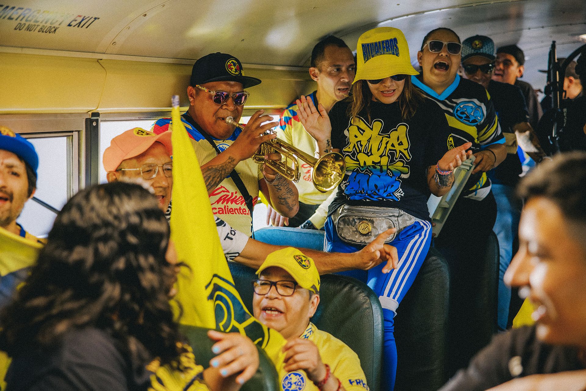 Group of people wearing colorful sports attire on a tightly packed bus, cheering and playing musical instruments. One person is playing a trumpet. Bright yellow and blue colors dominate their clothing.