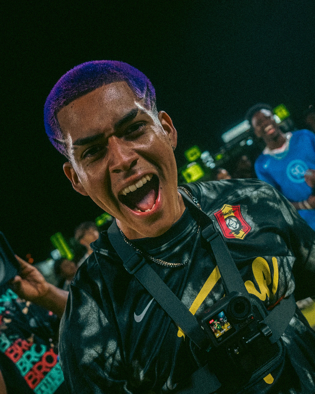 Person with purple hair wearing a black shirt and chest-mounted camera shouts energetically at an outdoor event, with people and bright lights in the background.