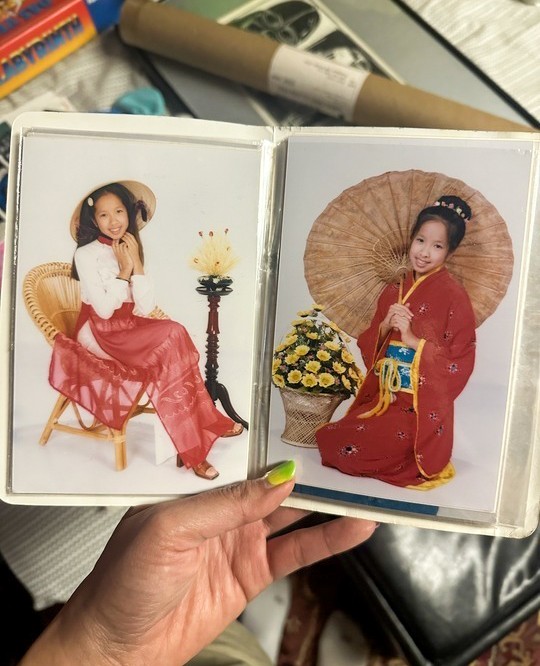 A hand holds open a photo album showing two portraits of a young girl in traditional Asian attire, each with a parasol and decorative props.