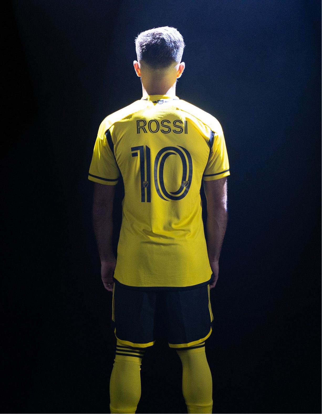 A soccer player stands facing away, wearing a yellow jersey with the name "Rossi" and the number 10, illuminated by a spotlight against a dark background.
