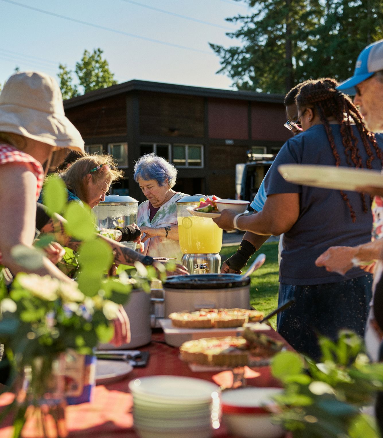A group of people serves themselves food and drinks from a buffet table outdoors on a sunny day, with a building and trees in the background.