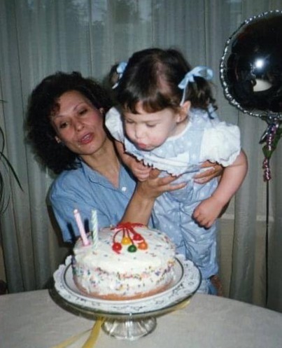 A woman holds a young girl as she blows out candles on a decorated birthday cake; a balloon is visible in the background.