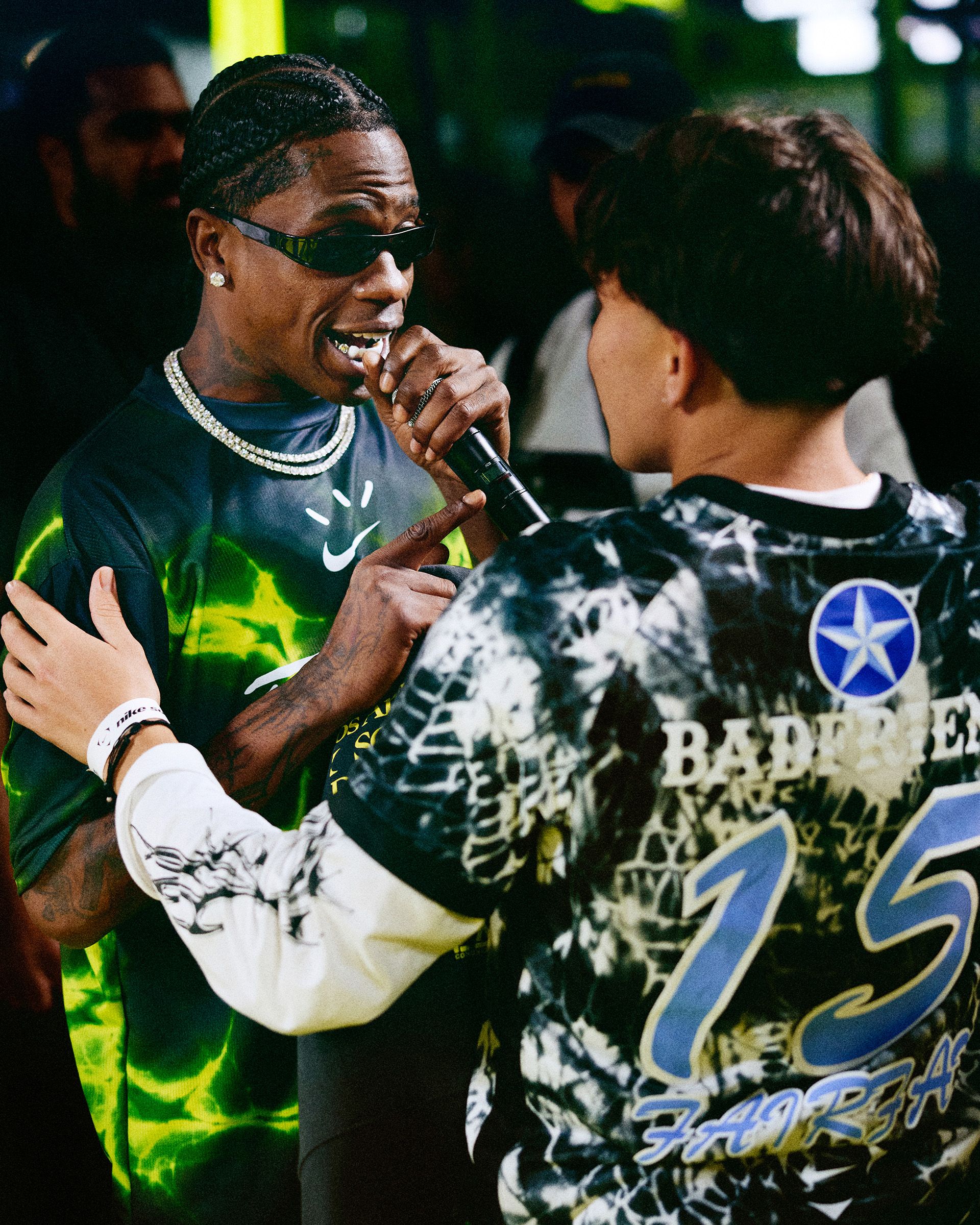 A man with braids and sunglasses holds a microphone, speaking to another person in a graphic shirt with "15" and "BAD BEAR" on the back, in a lively indoor setting.