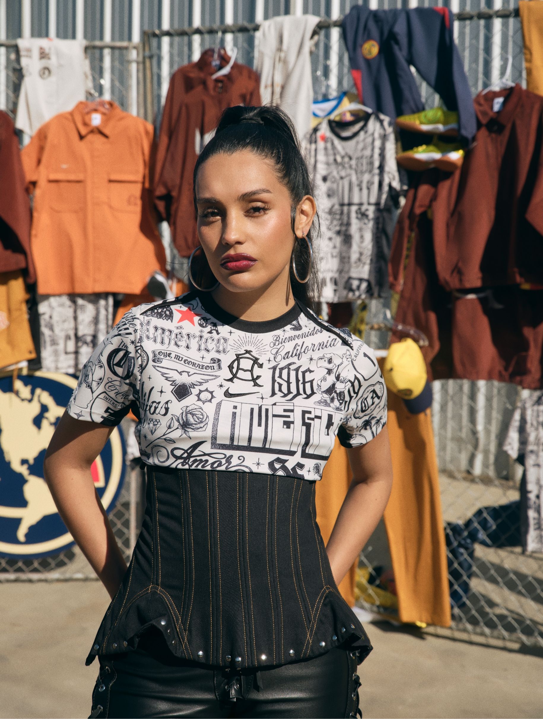 A woman stands outdoors in front of a fence displaying various shirts and jackets. She has long dark hair tied back, wears hoop earrings, and is dressed in a printed shirt and corset-style top.