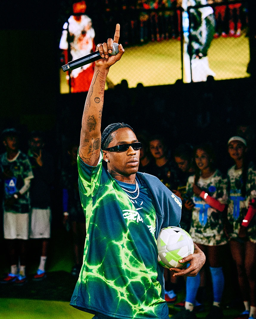 Man in a neon green sports jersey holds a soccer ball and microphone, raising one arm, with audience and large screen in the background.