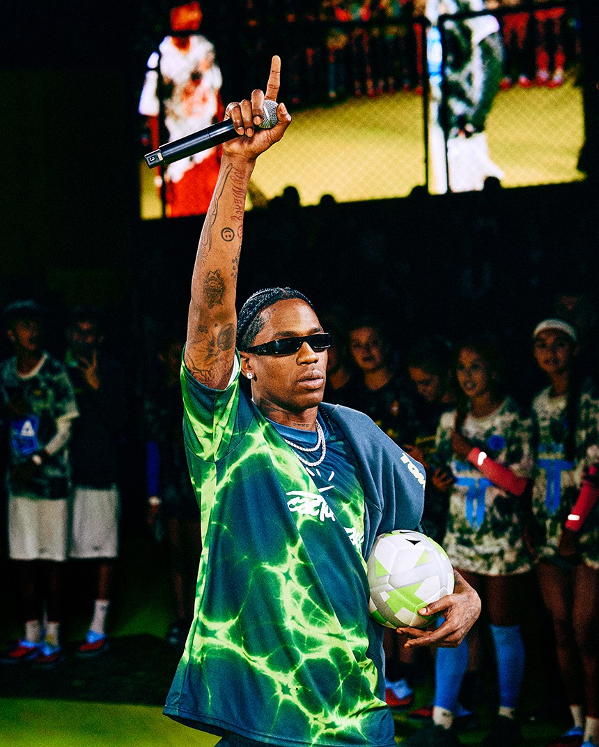 Man in a neon green sports jersey holds a soccer ball and microphone, raising one arm, with audience and large screen in the background.