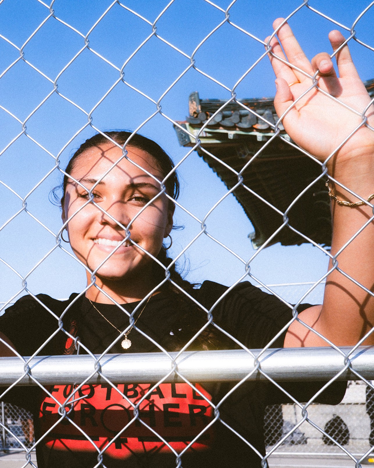 A person smiles while standing behind a chain-link fence, raising one hand. There is a building with a decorative roof in the background under a clear blue sky.