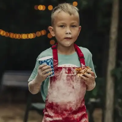 A young boy with flour on his face and red apron holds a paper cup and a slice of pizza outdoors at night; string lights are visible in the background.