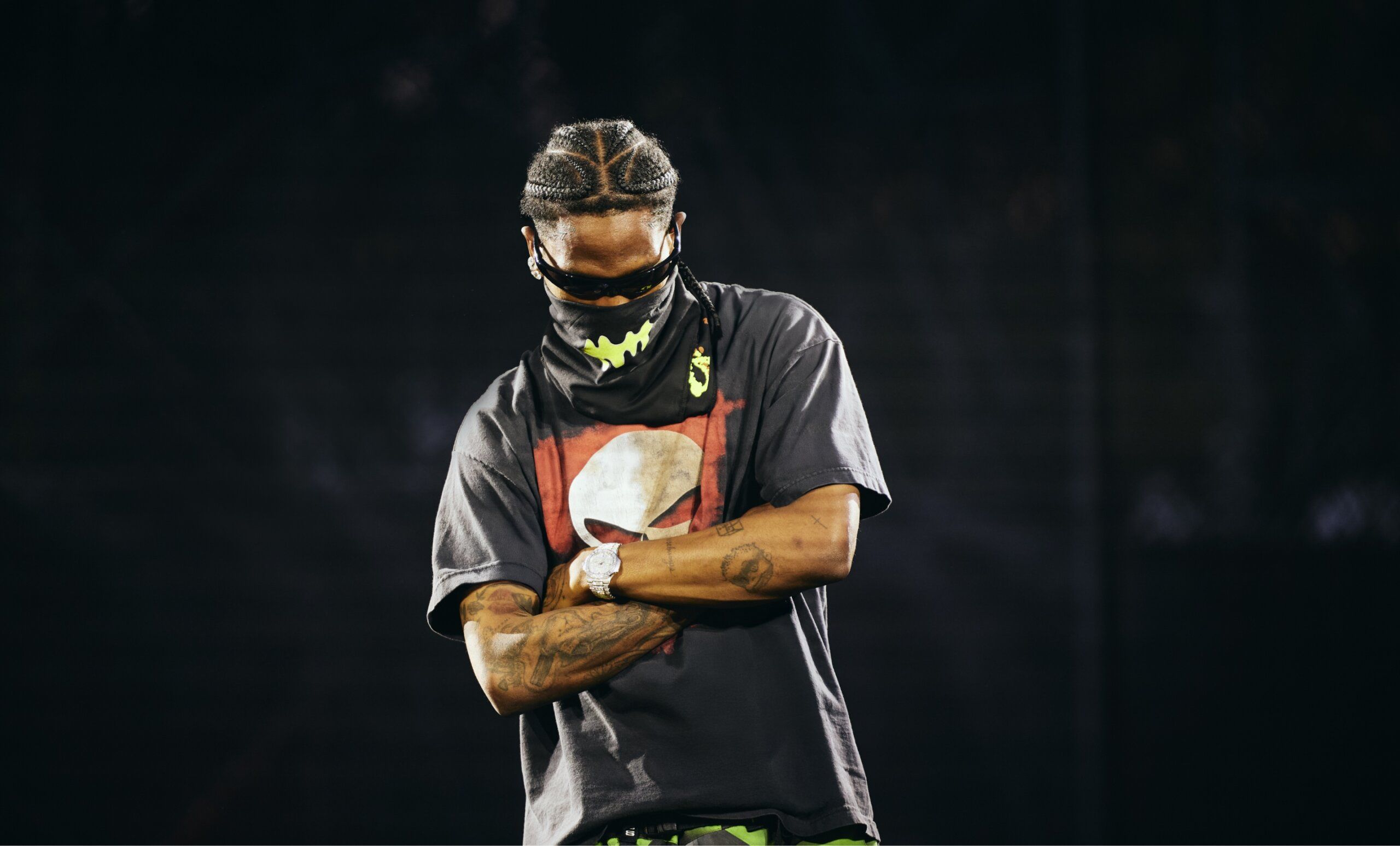 A person with braided hair, wearing a face mask, sunglasses, and a t-shirt with a red and white graphic, stands on stage with arms crossed.