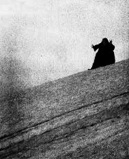 A solitary figure in dark clothing stands on a sloped, rocky surface against a grainy, overcast sky.