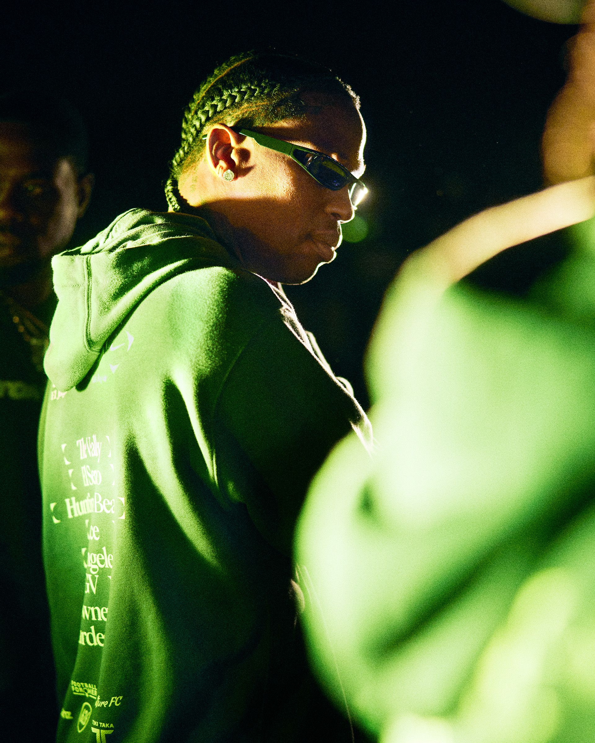 Person in a dark green hoodie and sunglasses stands under moody lighting, facing sideways, with braided hair; another figure is partially visible in the foreground.