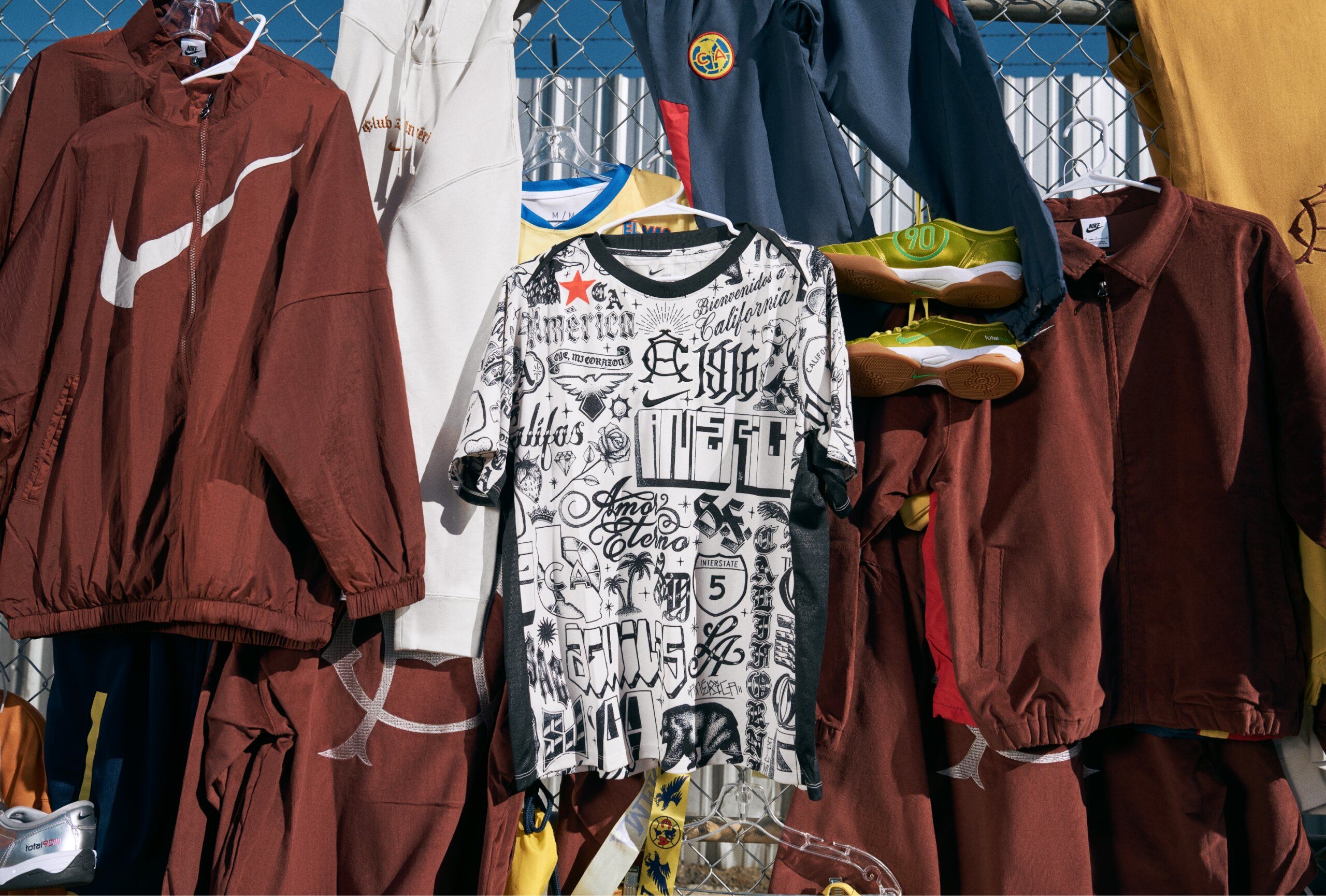 A white and black graphic T-shirt hangs on a fence, surrounded by brown, yellow, and blue athletic clothing and sneakers displayed outdoors.