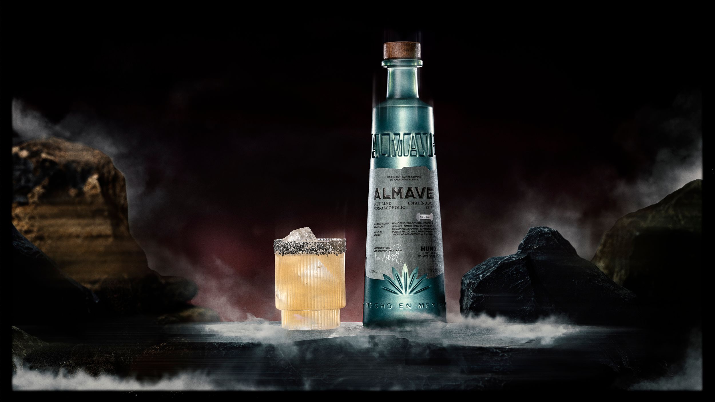 A tall bottle labeled "Almave" stands next to a glass of cocktail with ice and a salted rim, set on a rocky surface with mist and dark background.