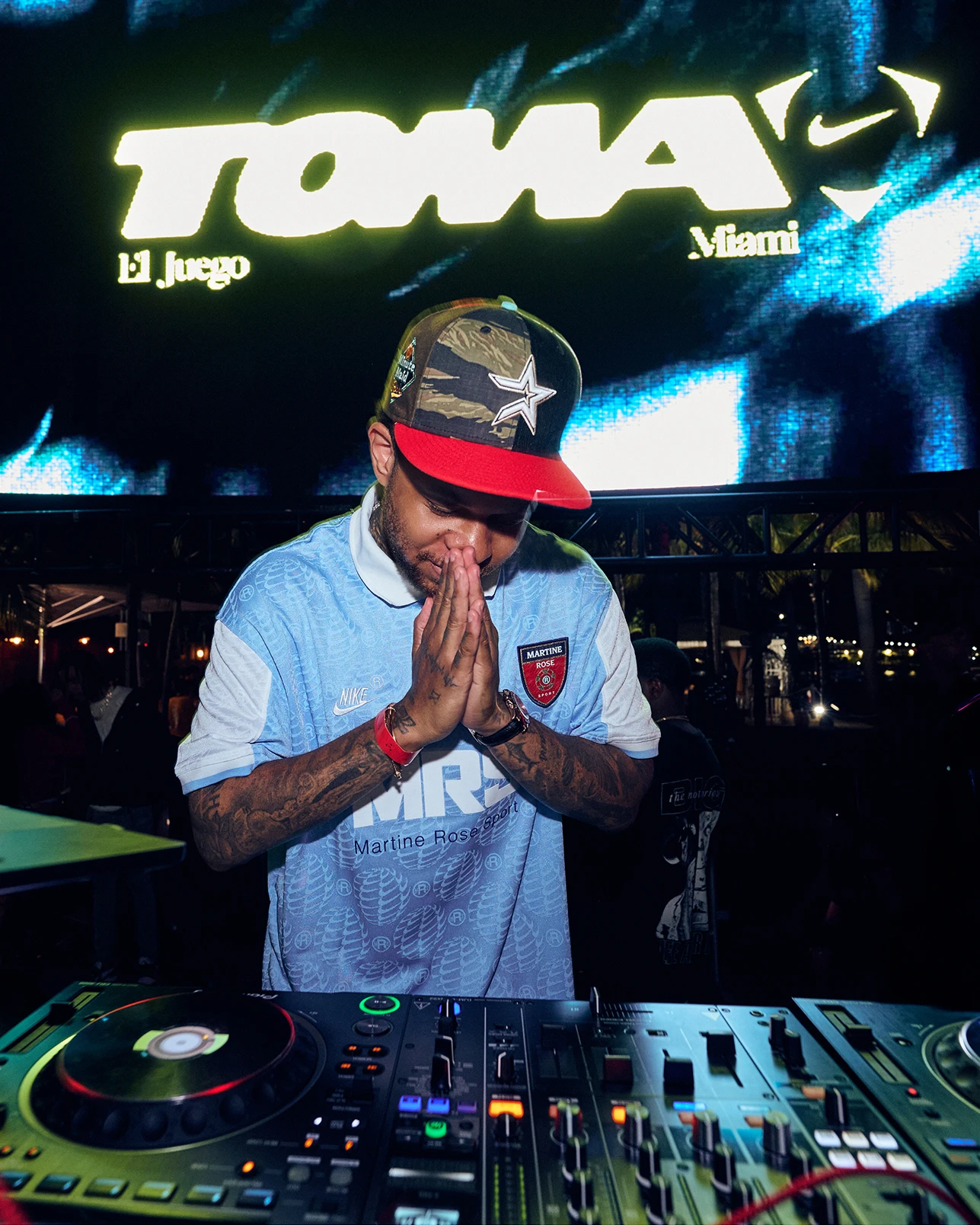 A DJ in a light blue soccer jersey and camo hat stands behind turntables, hands together in front of a large screen reading “TOMA.”.