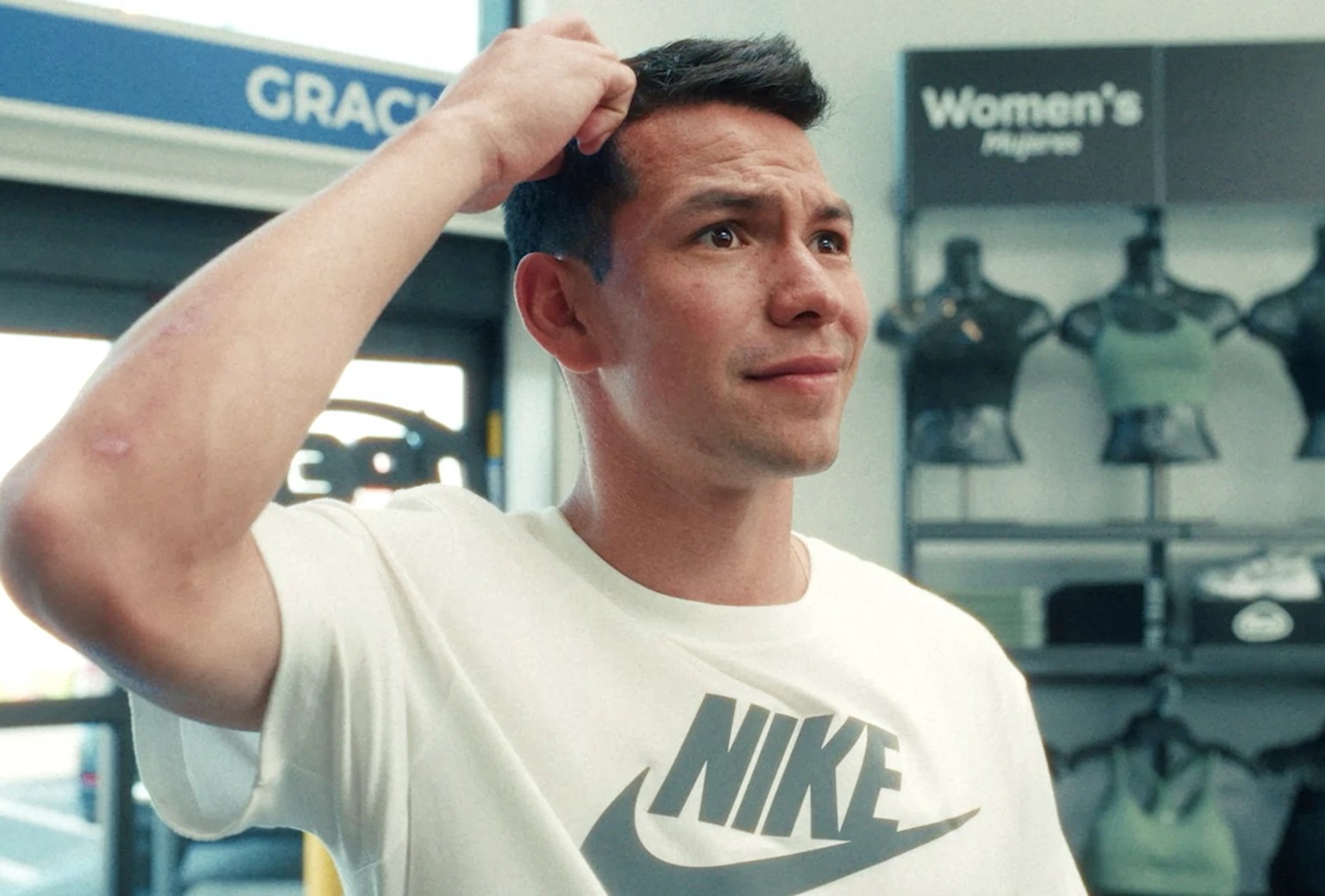 A man wearing a white Nike t-shirt stands indoors, scratching his head with a puzzled expression. Women's activewear is displayed in the background.