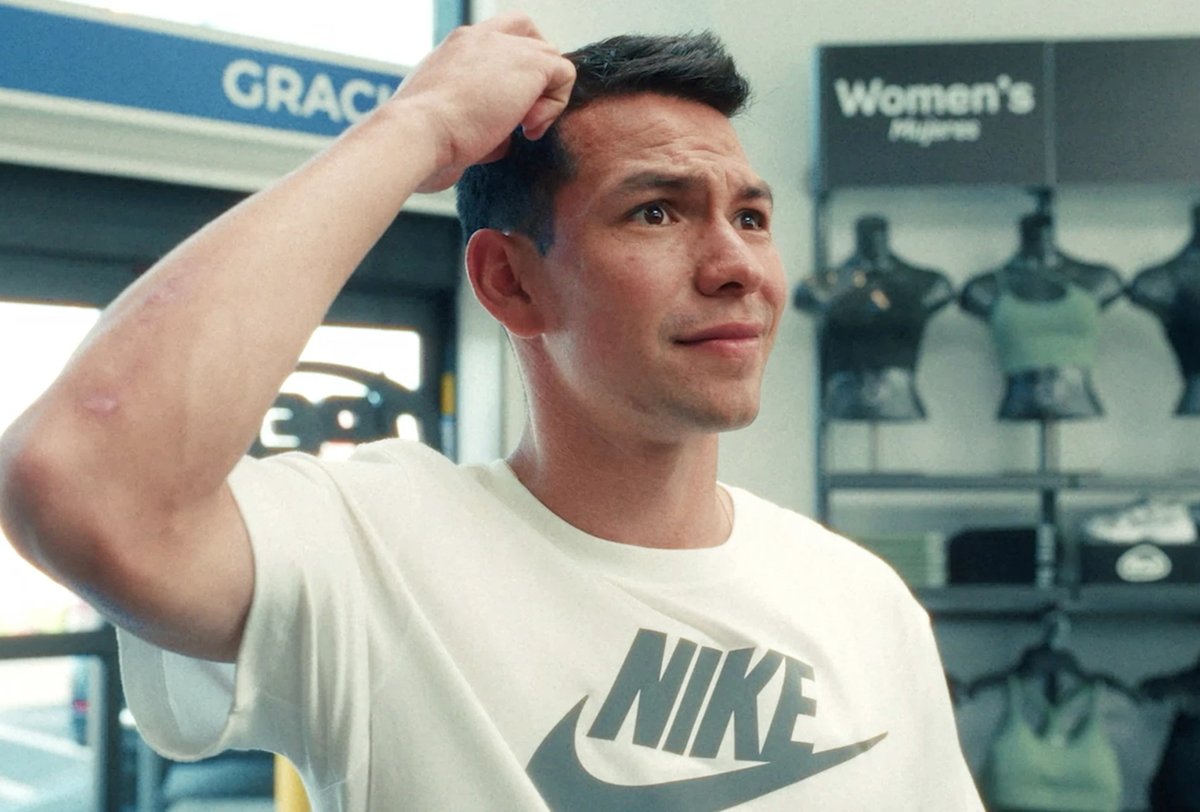 A man wearing a white Nike t-shirt stands indoors, scratching his head with a puzzled expression. Women's activewear is displayed in the background.