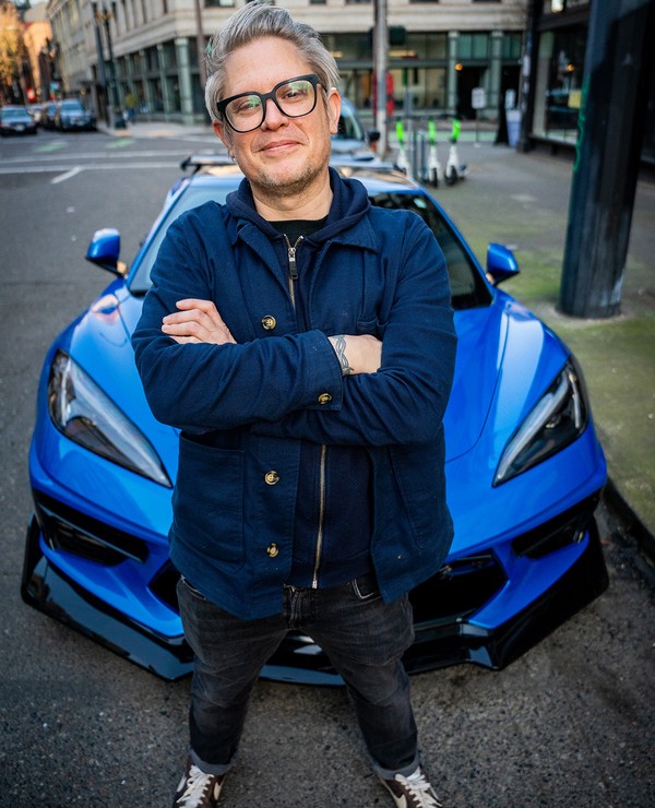 Person with glasses and gray hair stands with crossed arms in front of a blue sports car on a city street.