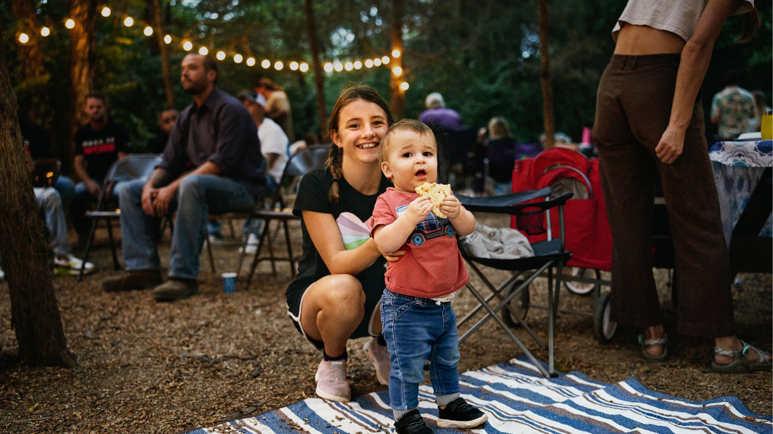 A young girl kneels behind a toddler eating a snack at an outdoor gathering, with string lights and people sitting in the background.