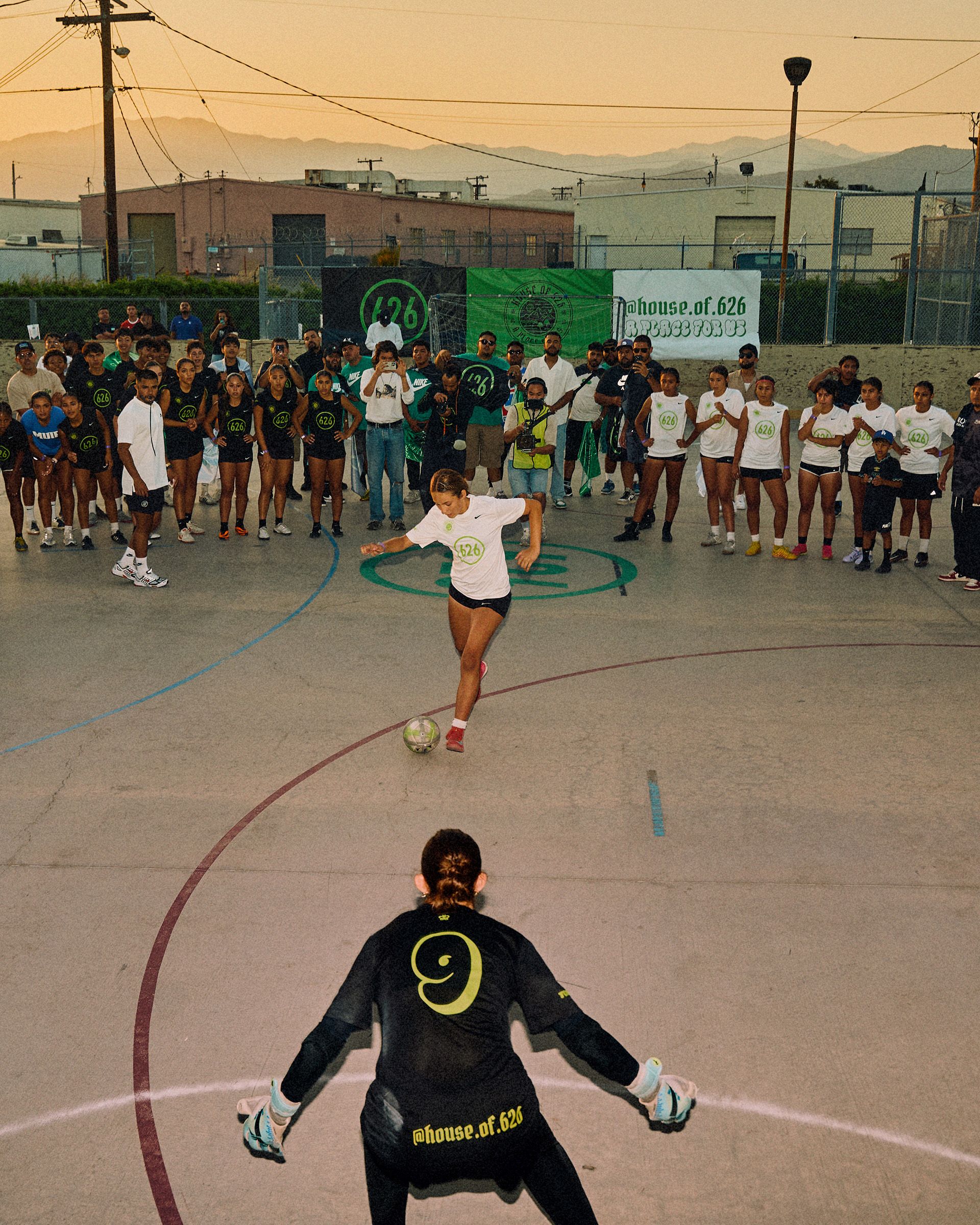 A soccer player prepares to kick a ball toward a goalie on an outdoor court, while a crowd watches from behind.