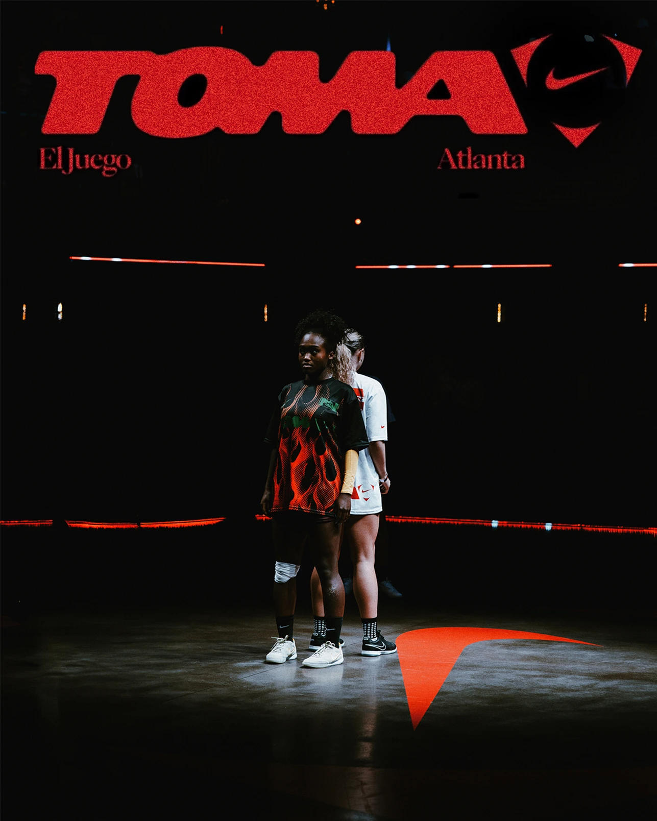 Two basketball players stand back to back on a dark court under a red “TOMA” sign, with spotlights highlighting them.