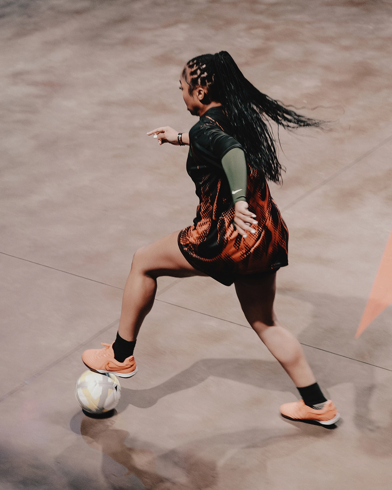 A person with long braids wearing a red and black outfit and orange sneakers is about to kick a soccer ball on an indoor court.