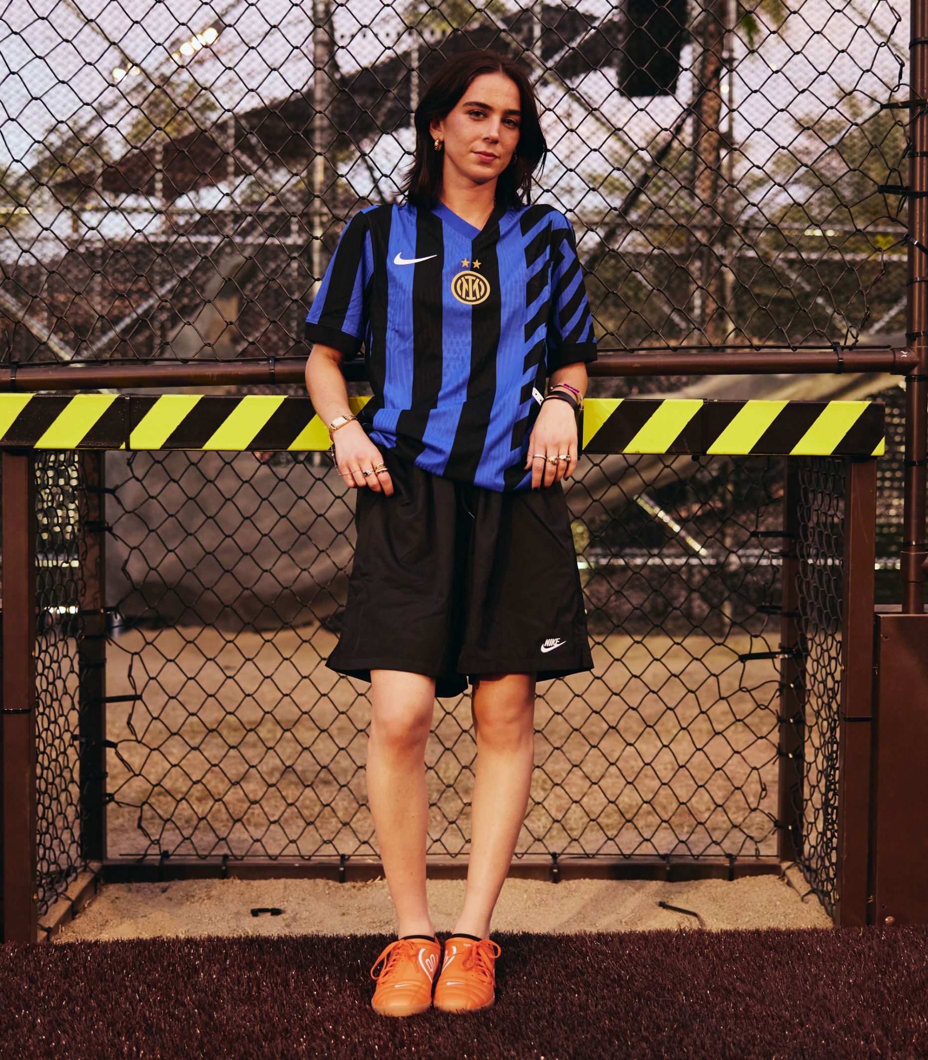 Person wearing a blue and black Inter Milan soccer jersey, black Nike shorts, and orange sneakers stands in front of a chain-link fence.